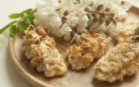 Fiori di acacia fritti, dorati e croccanti, su un piatto di legno, con grappoli di fiori freschi sullo sfondo.