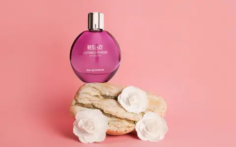 Różowa butelka perfum JFenzi Mon Amie Metamorphoze Women na kamieniu z białymi kwiatami.