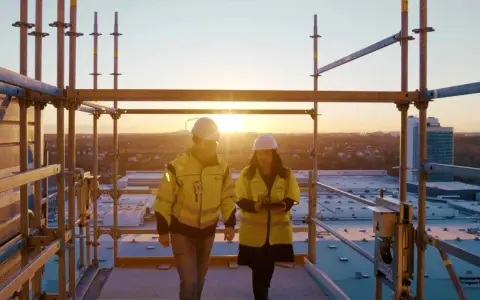 Två byggarbetare i varselkläder inspekterar ett bygge vid solnedgången. De diskuterar säkerhetslösningar, som brandklass EI60.