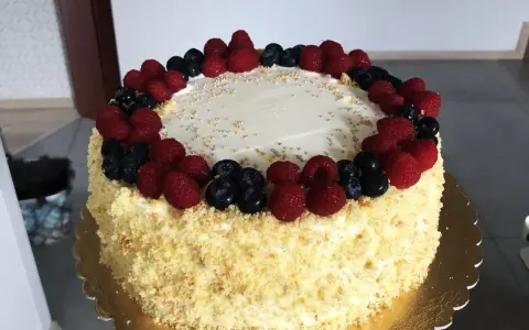Pyszny tort śmietankowo-malinowy z mascarpone, ozdobiony świeżymi malinami i borówkami, posypany złotymi i srebrnymi kuleczkami.