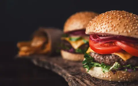 Dwa soczyste burgery z serem, pomidorem i czerwoną cebulą na drewnianej desce, obok frytki.