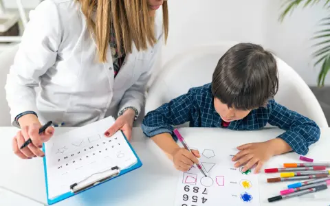 Bambino impegnato in un test per l'autismo, colora forme geometriche guidato da un'insegnante.