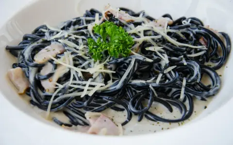 Czarny makaron spaghetti w kremowym sosie, posypany startym parmezanem i udekorowany gałązką natki pietruszki.