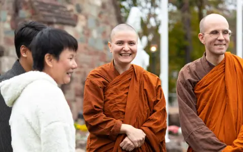 A smiling Buddhist monk in orange robes stands with others outside a church.