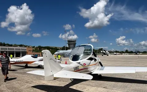 Zwei ultraleichtflugzeug modelle stehen auf dem Rollfeld. Im Hintergrund ein Kontrollturm und weitere Flugzeuge.