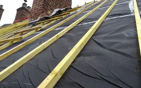 Charpente en bois et membrane noire sous une **ite toiture** en cours de construction, avec des cheminées en briques en arrière-plan.