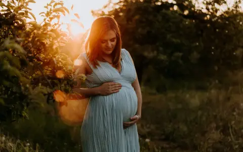 Piękne zdjęcia w ciąży. Kobieta w błękitnej sukience trzyma dłonie na brzuchu, otoczona ciepłym światłem zachodzącego słońca.