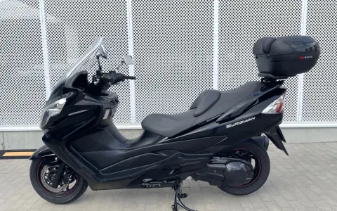 Czarny skuter Suzuki Burgman 400 z kufrem SH46 na tle perforowanej białej ściany.
