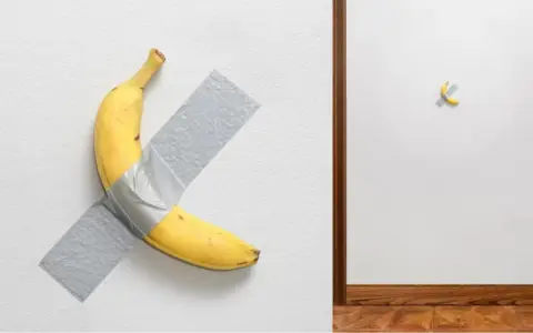 Un'opera d'arte banana, una gialla banana attaccata al muro con nastro adesivo grigio. A destra, un'altra banana, più piccola, fissata con nastro adesivo.
