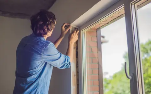 Un homme ajuste le joint d'une nouvelle pose fenêtre PVC. Le soleil brille à travers la vitre.