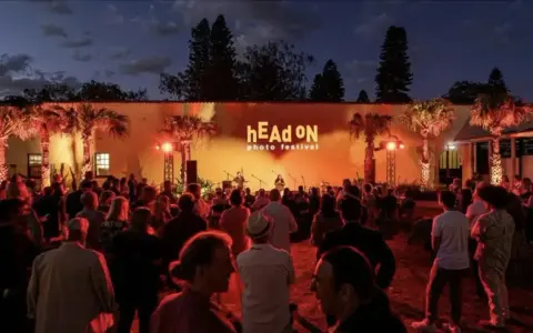 Crowd gathered at the hEAd oN photo festival, enjoying a performance under palm trees lit by warm orange lights.