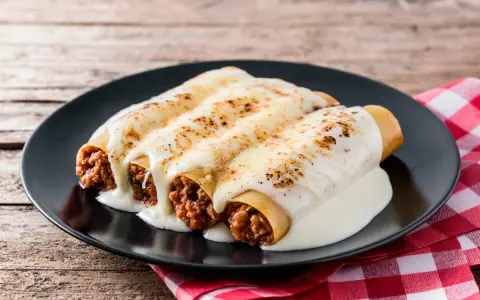 Canelones de carne picada cubiertos de queso fundido, servidos en un plato negro sobre una mesa de madera rústica.