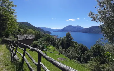 Sentiero del Viandante tappe: un casolare in pietra con tetto in ardesia e un recinto in legno rustico si affacciano su un lago blu intenso circondato da montagne verdi.