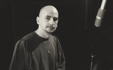Marek Pędziwiatr, z charakterystyczną łysą głową i wąsikiem, w ciemnej bluzie i z naszyjnikiem, przygotowuje się do nagrania.