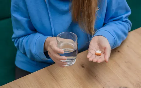 Kobieta w niebieskiej bluzie trzyma szklankę wody i kilka tabletek na dłoni.