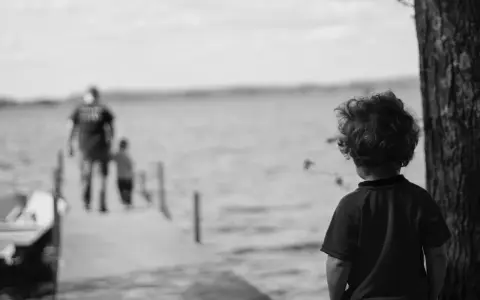 Bambino con capelli ricci guarda verso un molo sfocato dove camminano due figure. Un senso di isolamento, quasi un attaccamento disorganizzato, aleggia nell'aria.