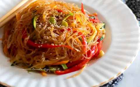 Un piatto di spaghetti di soia con verdure colorate, peperoni rossi, zucchine e carote, guarnito con semi di sesamo.