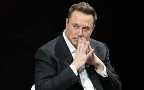 Elon Musk, znany wizjoner i przedsiębiorca, w czarnym garniturze, z dłońmi złożonymi przed twarzą.