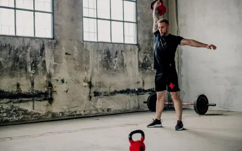 Mężczyzna wykonuje ćwiczenia z kettlebell, idealne dla początkujących. W tle sztanga i surowe wnętrze.