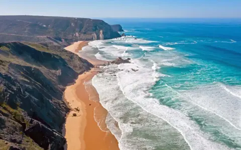 Atemberaubende Küstenlandschaft im Distrikt Faro, Portugal. Steile Klippen treffen auf einen goldenen Sandstrand und das türkisfarbene Meer mit tosenden Wellen.