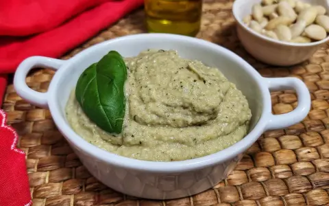 Pesto di melanzane cremoso in una ciotola bianca, guarnito con una foglia di basilico. Sullo sfondo, mandorle e olio d'oliva.