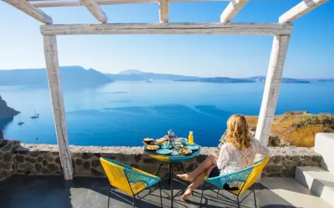 Mujer desayunando en balcón con vista al mar azul y montañas.