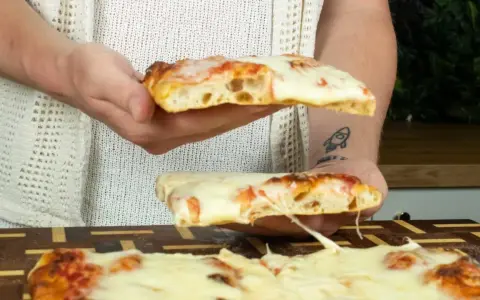 Due fette di pizza in teglia, con mozzarella filante e crosta dorata, vengono sollevate da mani.