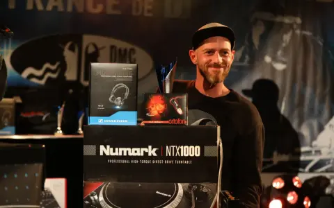 Un DJ souriant pose avec des prix et du matériel, dont des casques Sennheiser et un tourne-disque Numark. Une liste DJ internationale est visible.