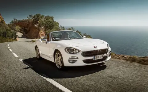 Biały Fiat 124 Spider sunie po krętej drodze nad morzem.