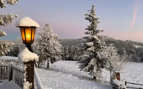 Zimowy krajobraz o zmierzchu. Ośnieżone drzewa i pola. W oddali widać zarys gór. Latarnia przy schronisku Orzeł zaprasza do środka.