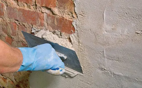 Hand mit blauer Handschuh glättet mit Kelle den **Sanierputz Keller** auf rotem Ziegelmauerwerk.