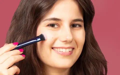 Una mujer sonríe mientras aprende como aplicar colorete con brocha, logrando un rubor natural en sus mejillas.