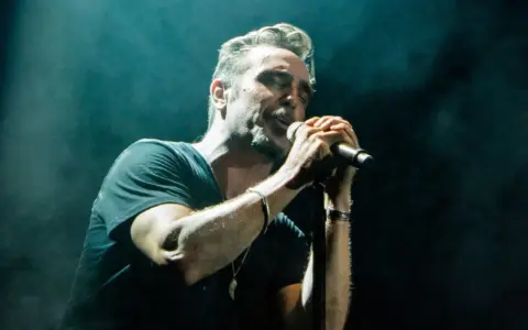 Un miembro de OBK canta apasionadamente en el escenario, iluminado por focos.