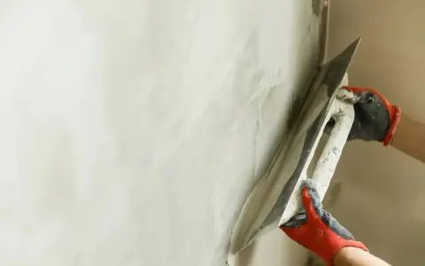 Ręce w rękawiczkach rozprowadzają gładź gipsową po ścianie za pomocą kielni.