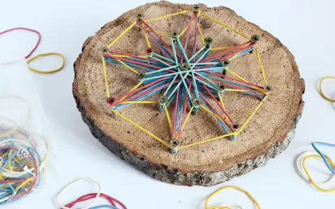 Bunte Gummibänder spannen sich zu einem Stern auf einer Baumscheibe. Ein tolles Bastelprojekt für Kinder, das Kreativität und Feinmotorik fördert.