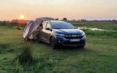 Dacia Jogger Camper rozłożony na biwaku o zachodzie słońca. Namiot połączony z autem, otoczony łąką i wodą.