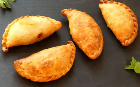 Cuatro empanadillas doradas y crujientes dispuestas sobre una superficie oscura, con hojas de menta a los lados.