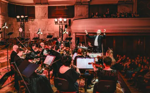 Dyrygent prowadzi orkiestrę filharmoniczną. Koncert trwa, a widzowie na balkonie wsłuchują się w muzykę.