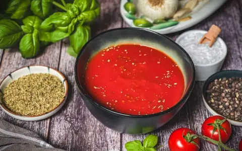 Schüssel mit leuchtend roter Pizza Sauce, umgeben von frischem Basilikum, Tomaten, Oregano, Salz und Pfeffer – wie in der Pizzeria.