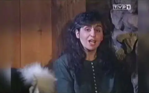 Kobieta z ciemnymi włosami, w zielonej bluzce, mówi do kamery. Chciałabym być tam gdzie ty, w tym przytulnym wnętrzu.