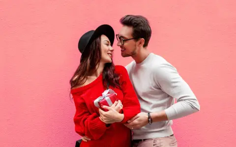 Para zakochanych na tle różowej ściany. Ona trzyma prezent, on obejmuje ją ramieniem. Mówi mu "I love you" po angielsku.