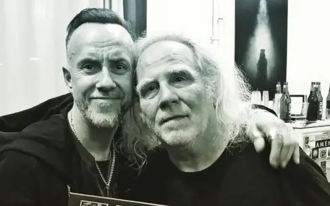 Dwóch mężczyzn, jeden z nich to Roman Kostrzewski, pozuje do zdjęcia. W tle widać butelki piwa i plakat.