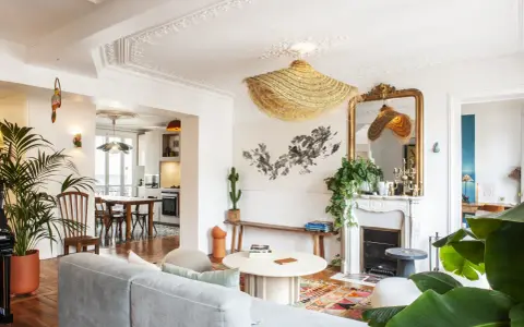 Salon au style classique revisité, avec un canapé gris, un tapis coloré, une cheminée ornée et des plantes luxuriantes.