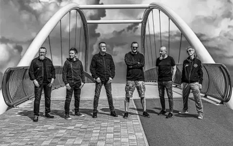 Sześciu mężczyzn, zespół Strachy na Lachy, pozuje na moście. Gotowi na koncerty, w rockowym stylu, pod burzliwym niebem.