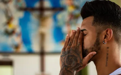 A man with tattoos prays before a crucifix, contemplating if is vaping a sin.