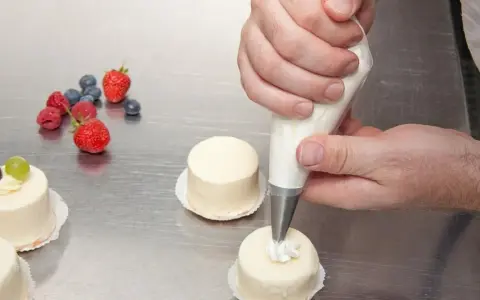 Man usa una manga pastelera para decorar pequeños pasteles. Frutas rojas y arándanos adornan la mesa.