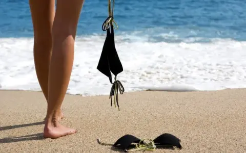 Nogi na piaszczystej plaży, obok czarny kostium kąpielowy. Fale obmywają brzeg. Idealne miejsce na naturystyczną plażę.