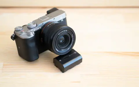 Srebrno-czarny aparat Sony a7c z obiektywem i zapasową baterią na drewnianym tle.