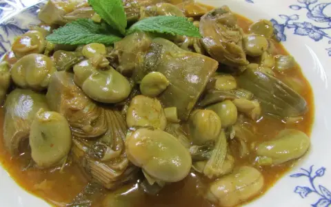 Plato de habas tiernas y trozos de alcachofa en una salsa dorada, decorado con hojas de menta fresca.