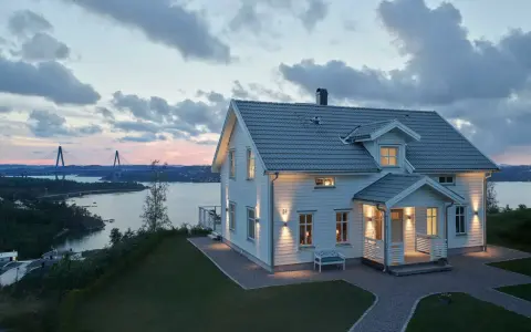 Vackert vitt hus med upplysta fönster och entré, perfekt hus entre inspiration. Vacker vy över vatten och bro i skymningen.